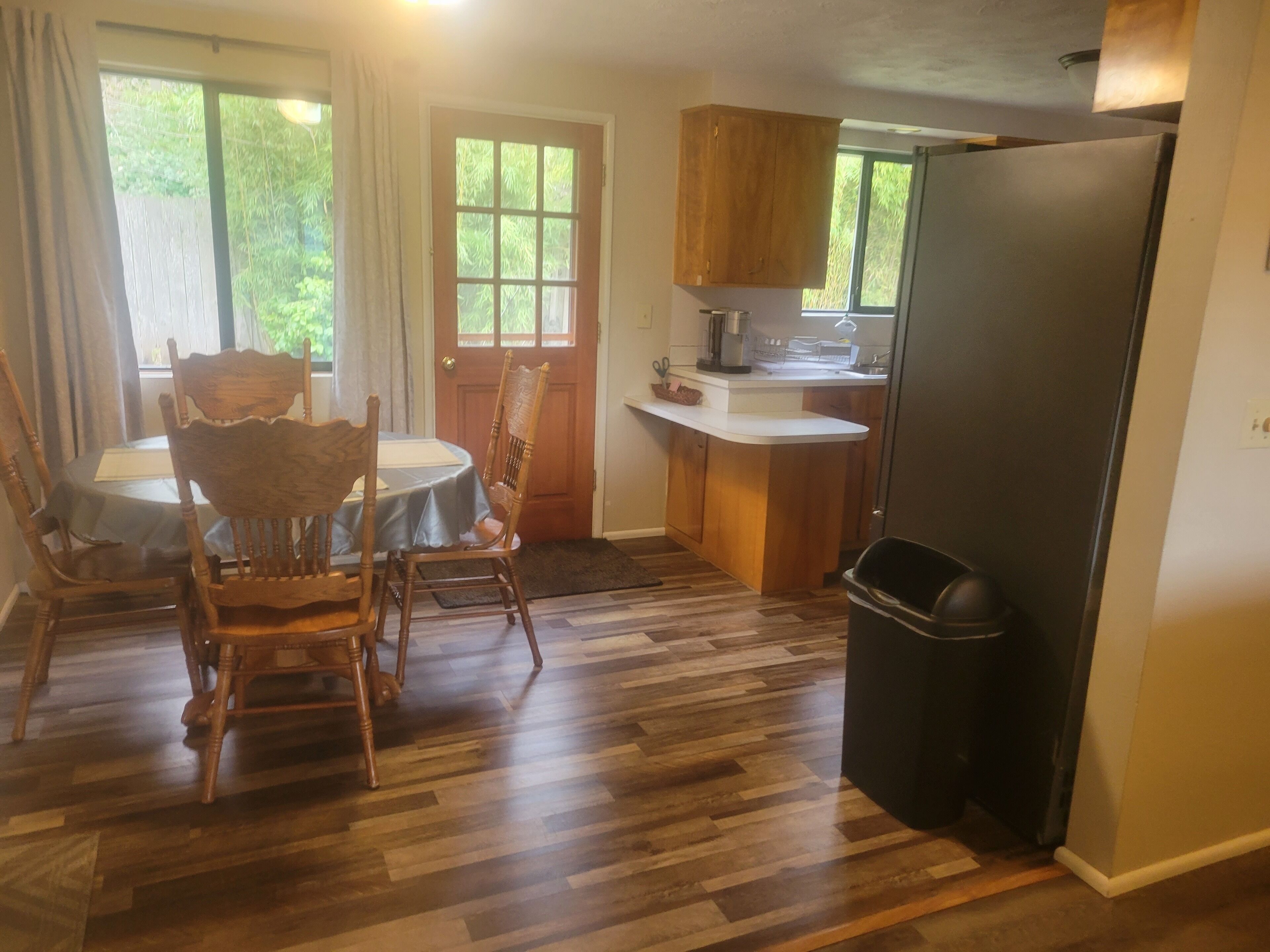 Un coin repas avec une table et des chaises, une cuisine avec des meubles en bois et un réfrigérateur, et une poubelle.