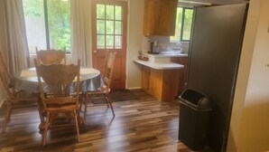 A dining area with a table and chairs, a kitchen with wooden cabinets and a refrigerator, and a trash bin. - South Hills Rose house (Eugene)