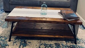 Un salon avec un canapé gris, deux coussins bleu foncé, une table basse en bois sur laquelle se trouvent une bouteille en verre et un livre, un tapis à motifs et une petite étagère avec une plante.