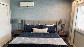 Une chambre à coucher avec une tête de lit capitonnée, un lit recouvert d’une courtepointe bleu foncé, deux tables de chevet avec des lampes et une fenêtre avec des stores.