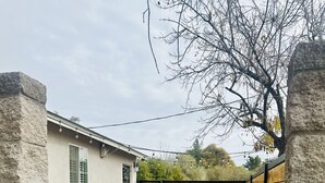 Une propriété résidentielle avec un chemin en béton menant à un portail en bois, une maison blanche dotée d’une fenêtre et un espace patio aménagé avec du mobilier d’extérieur.
