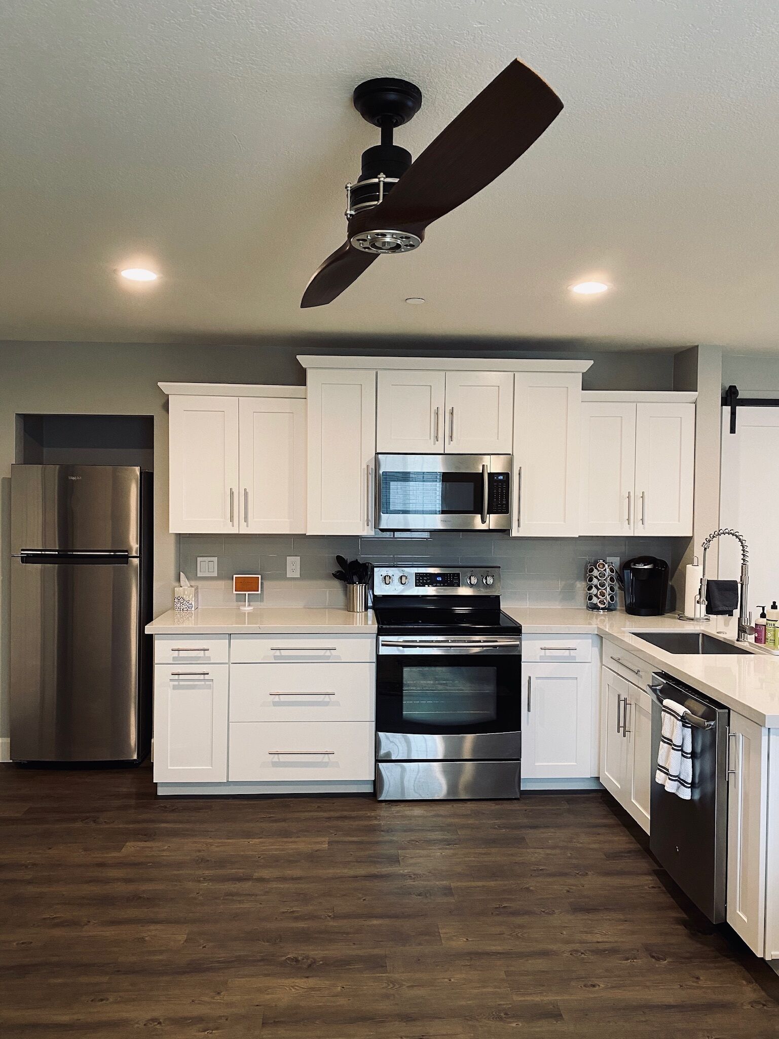 Una cocina moderna con armarios blancos, electrodomésticos de acero inoxidable, un ventilador de techo negro y suelo de madera.