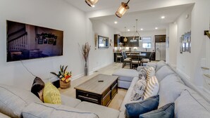 Uma sala de estar espaçosa com uma televisão de tela grande, um conjunto de sofás, uma mesa de centro e uma área de jantar com cadeiras, além de uma cozinha ao fundo.