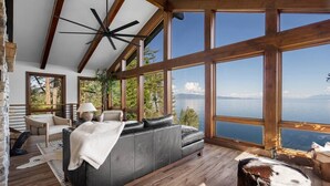 A spacious living room with a large black leather sofa, wooden flooring, and a view of a lake through large windows. - Modern Mountain Lake House - 3 bedrooms + Master Loft (Polson)
