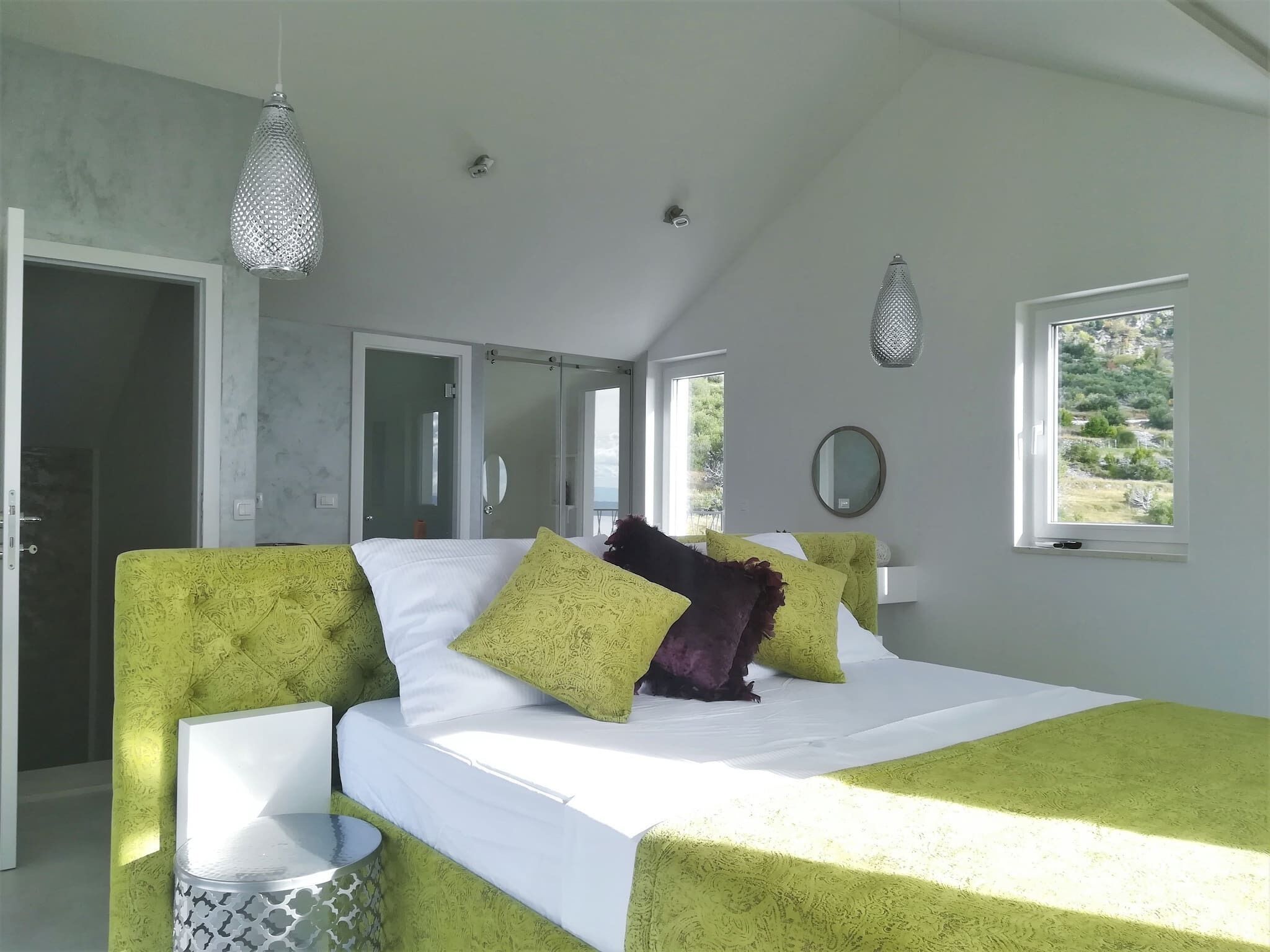Une chambre à coucher avec un lit, des meubles recouverts d’un tissu vert, un luminaire suspendu, une fenêtre donnant sur un paysage et un miroir au mur.