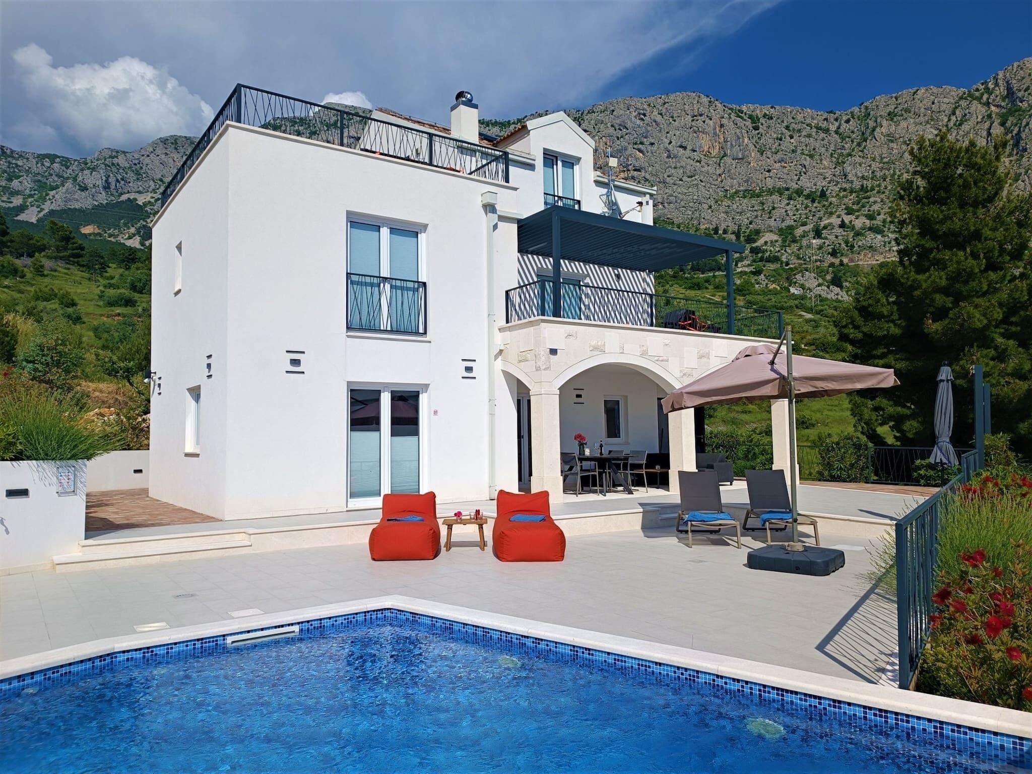 A modern two-story house with a swimming pool, outdoor seating, and a view of a mountainous landscape.