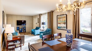 A living room with a TV, blue armchairs, a wooden coffee table, and a dining area with a vase and placemats. - Stay in Franklin - Close to downtown - fire pit & grill (Franklin)