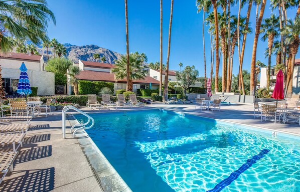 A swimming pool with lounge chairs and umbrellas, surrounded by palm trees and mountains in the background.