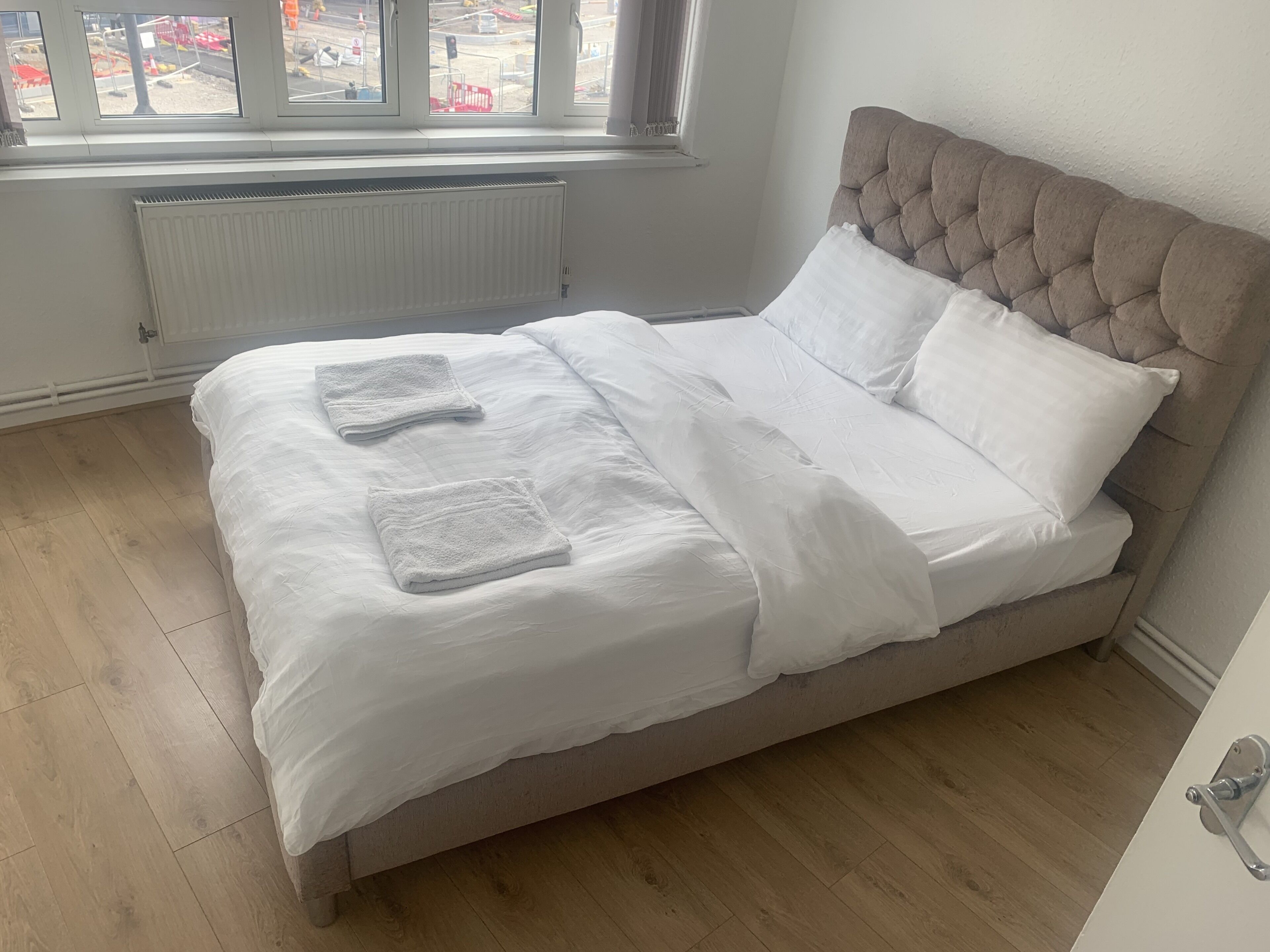 Un lit bien fait, avec du linge de lit blanc et deux serviettes pliées, une tête de lit capitonnée et, par la fenêtre, une vue sur un chantier de construction.