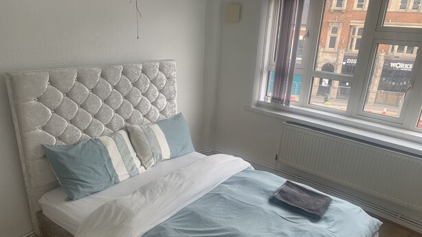 A neatly made bed with a tufted headboard, a window with a view of buildings, and a radiator below. - Birmingham city centre apartment  (Birmingham)