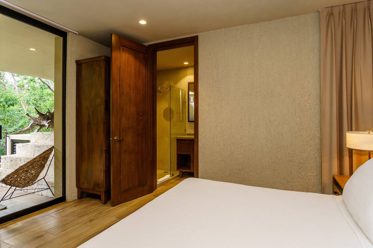 Une chambre avec une armoire en bois, une porte en verre donnant sur une salle de bain, un lit et une chaise près d’une porte en verre.