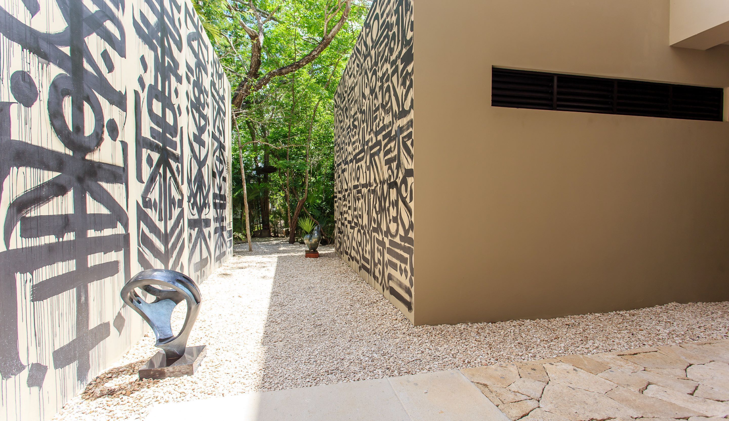 Un étroit sentier bordé de murs aux motifs décoratifs, une sculpture en métal et un chemin de gravier.