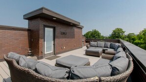 A rooftop patio with a brick wall, a door, and a seating area with wicker furniture and cushions.
