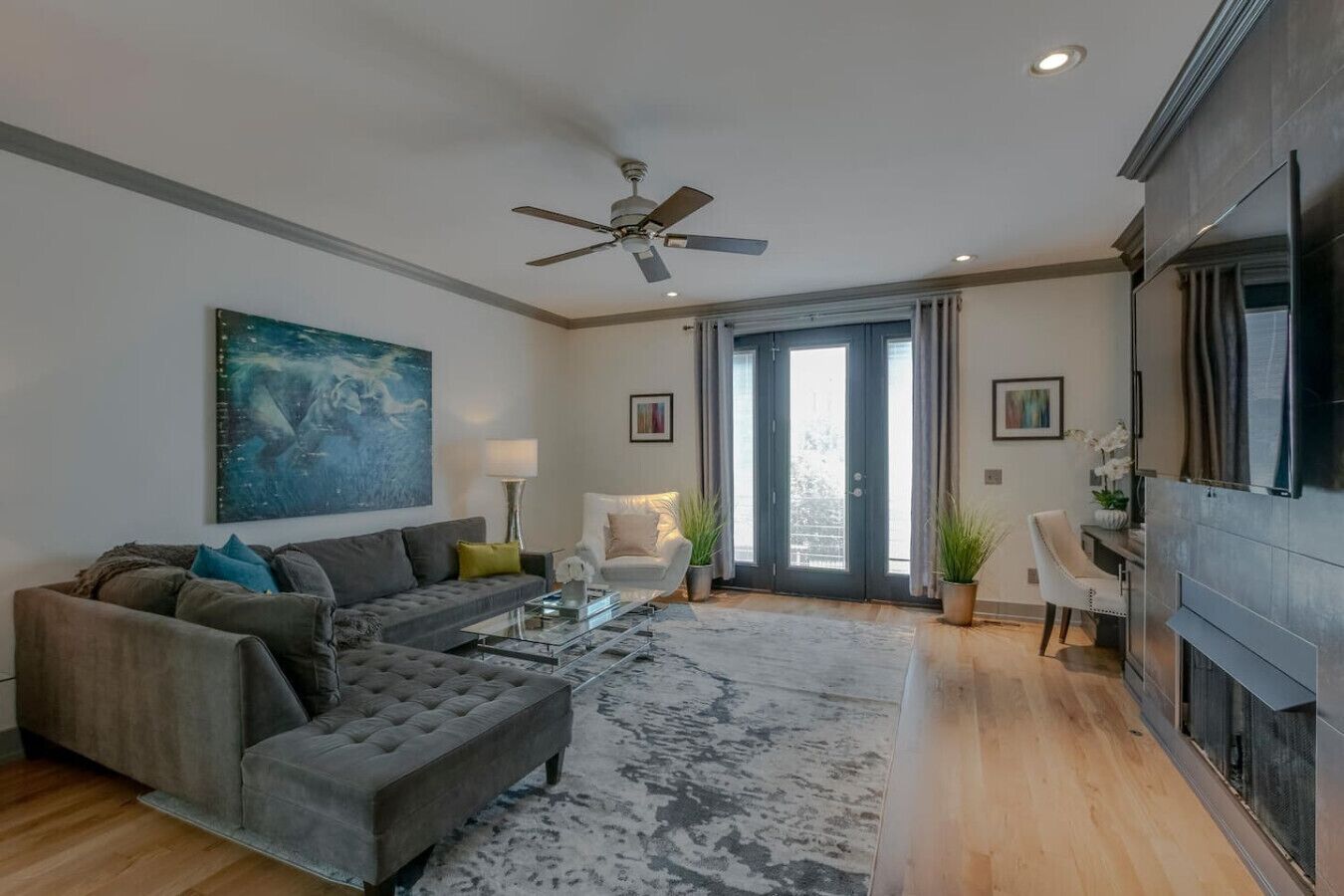 A modern living room with a grey sectional sofa, a coffee table, and a ceiling fan. There is a large painting on the wall and a fireplace on the right.