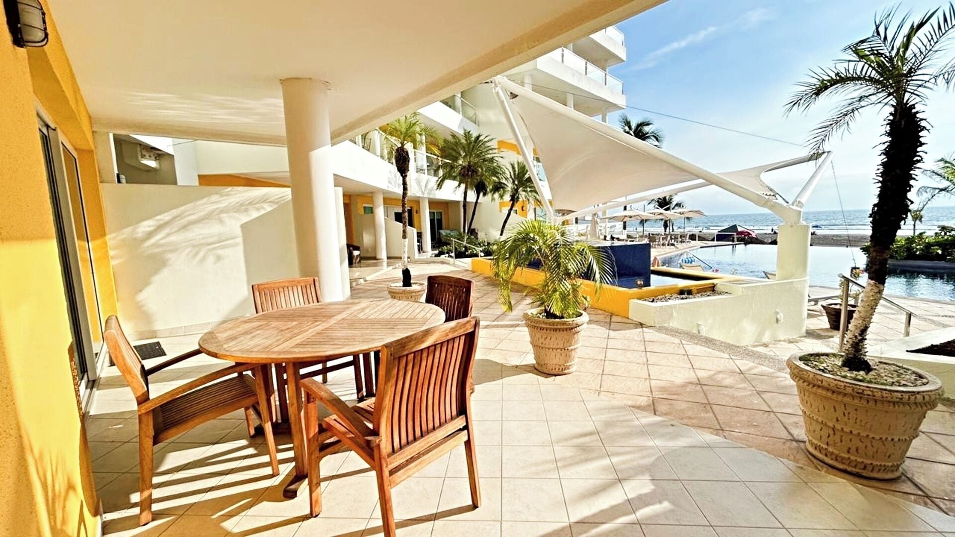 An outdoor patio area with a wooden table and chairs, potted plants, and a view of the pool and ocean.