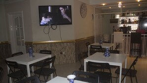 A restaurant with white tables and black chairs, a TV mounted on the wall, and a bar area in the background. - Casa Suit Capir (Malabo)