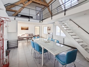 Een modern interieur met een eethoek met een glazen tafel en blauwe stoelen, een trap en een woonkamer met een bank en kunstwerken.