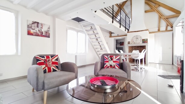 Ein großzügiger Wohnbereich im modernen Stil, mit einer Couch und Sesseln mit Union-Jack-Kissen, einem Glastisch und einer Treppe, die in einen oberen Bereich führt.
