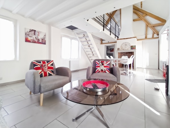 Een ruime woonruimte met een modern design, met een bank en fauteuil met Union Jack kussens, een glazen salontafel, en een trap naar een hoger gelegen verdieping.