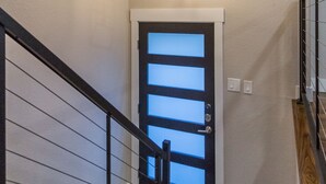 Uma escadaria com corrimão de madeira, uma porta preta com painéis de vidro jateado e um lustre geométrico acima.