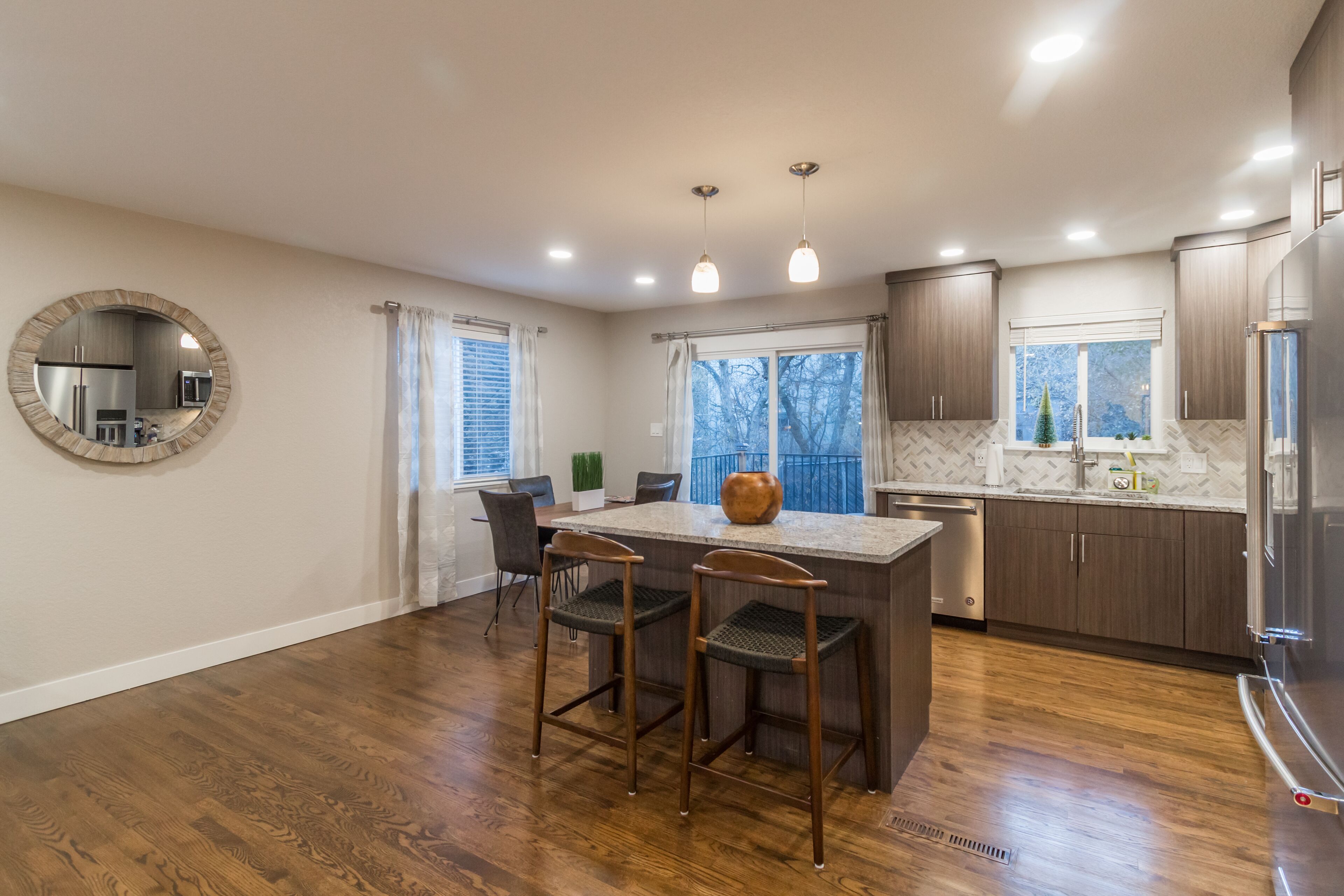 A modern kitchen with wooden flooring, a central island with bar stools, a round mirror, and a view of the outdoors through large windows.