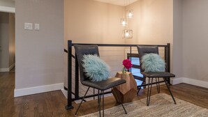Uma área de espera moderna com duas cadeiras pretas, uma mesinha de madeira com um vaso de flores e uma luminária com design geométrico.