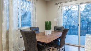 Uma área de refeições com mesa de madeira e quatro cadeiras, uma planta em vaso e vista para uma varanda com corrimão.