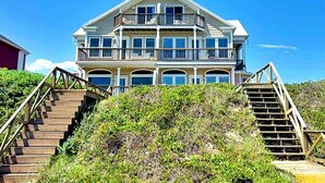 Una propiedad frente al mar de varios pisos, con un balcón en cada nivel, escaleras de madera que conducen a la entrada y una vista despejada del océano.