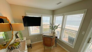 Una habitación con escritorio, silla y televisor en la pared. Hay ventanas con persianas, una mesita con un florero y una cesta en el suelo.