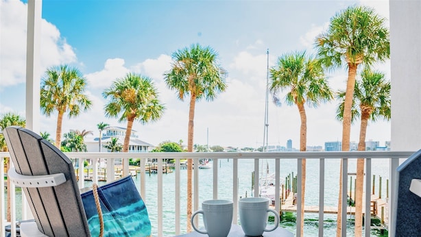 A balcony with a view of the water, palm trees, and a building in the background. - 2 Side-by-Side Waterfront Condos • 4BR/4BA • Walk to Beach (Clearwater)