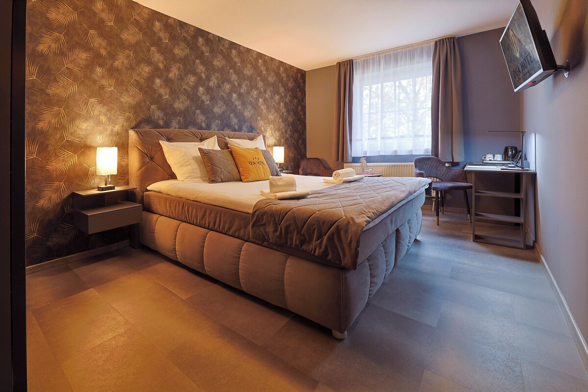 Habitación de hotel con una cama grande, un escritorio y una silla. Hay una ventana con cortinas, una lámpara en la mesita de noche y una pared con un diseño de hojas.