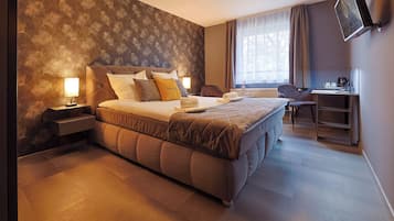 A hotel room with a large bed, a desk, and a chair. There is a window with curtains, a lamp on the nightstand, and a wall with a leaf pattern.