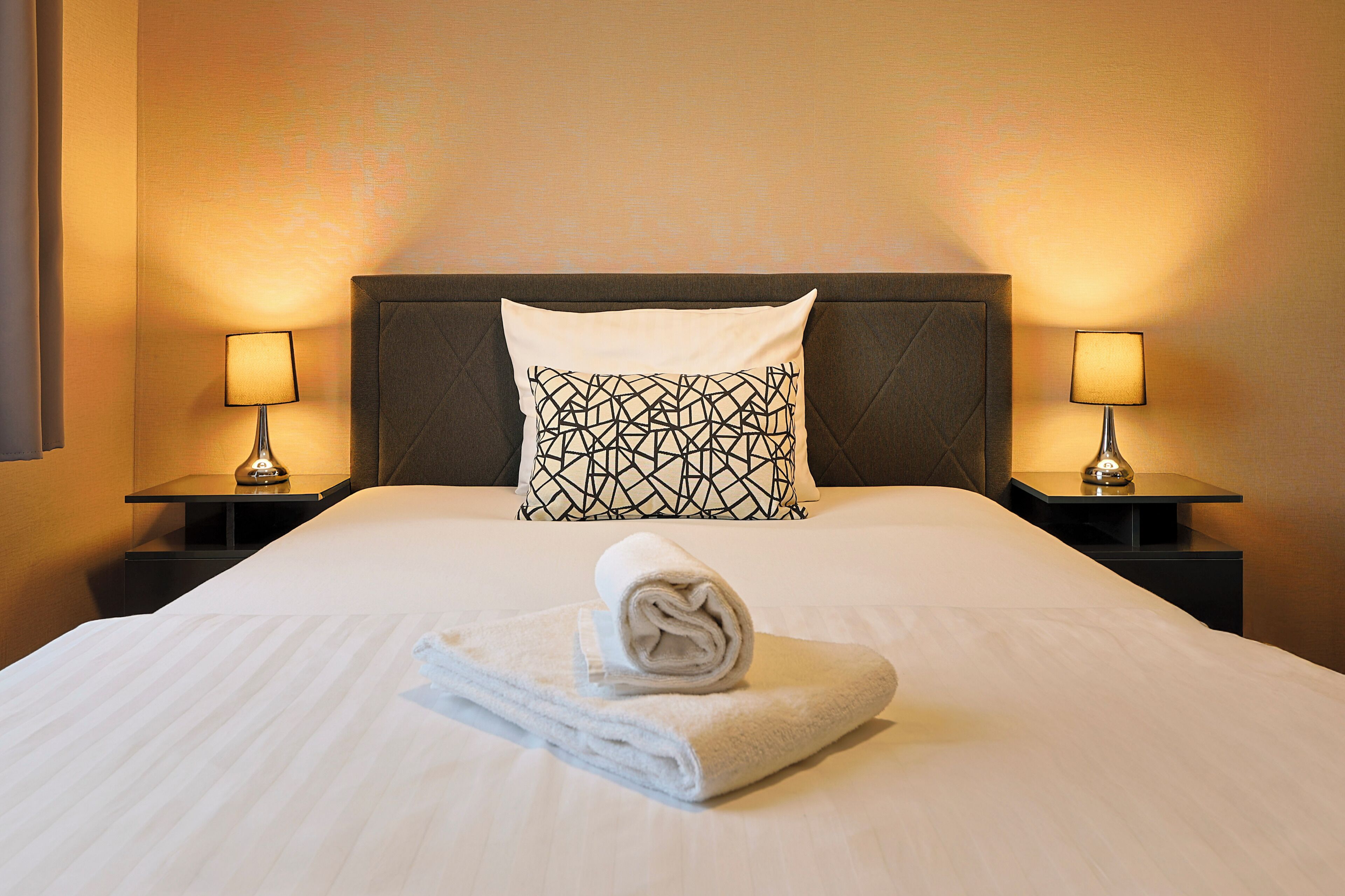 A neatly made bed with a rolled towel, two bedside lamps, and a pillow with a geometric pattern.