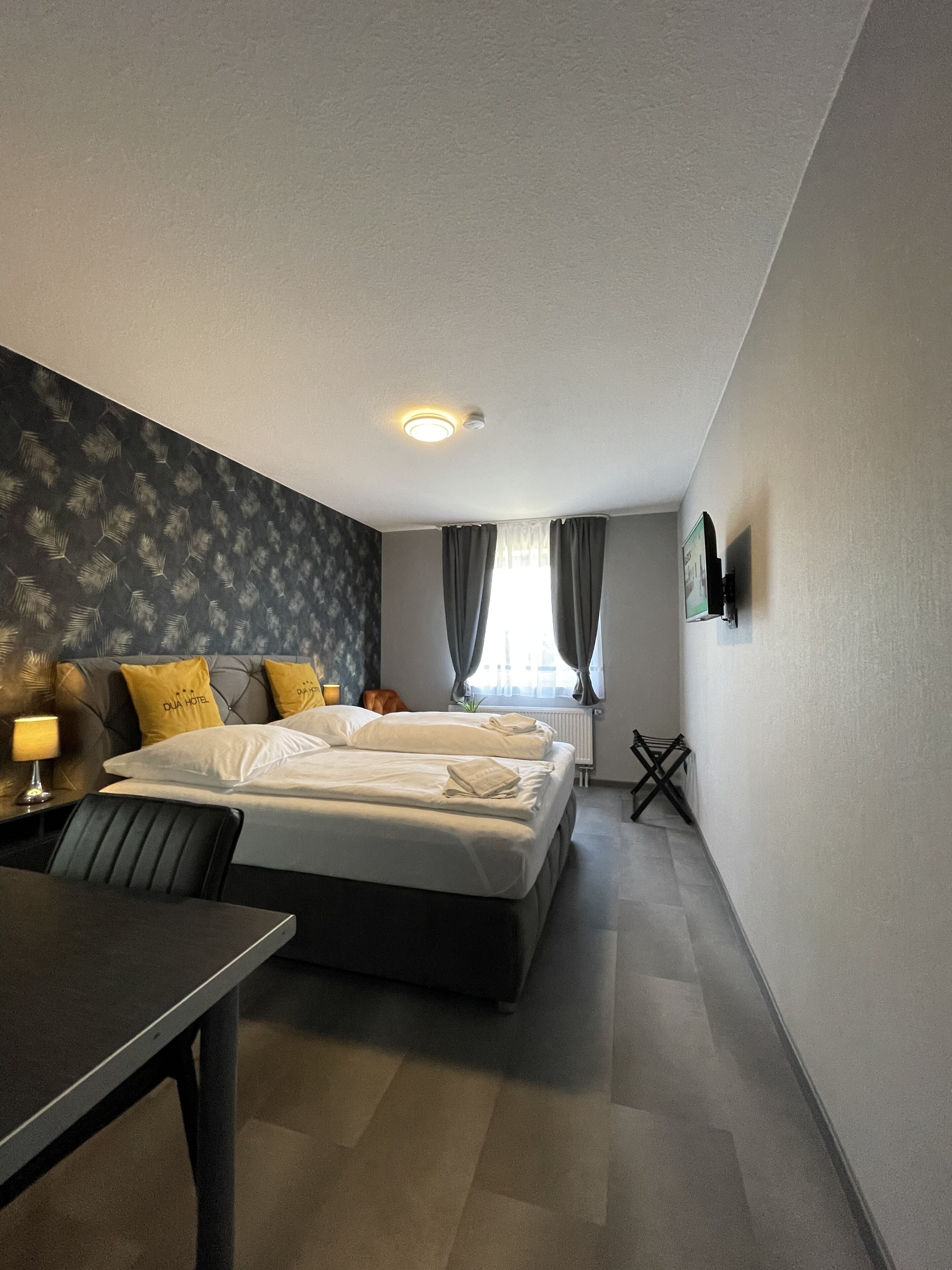 A hotel room with a bed, a desk, and a chair. There is a window with curtains, a radiator, and a wall-mounted TV.