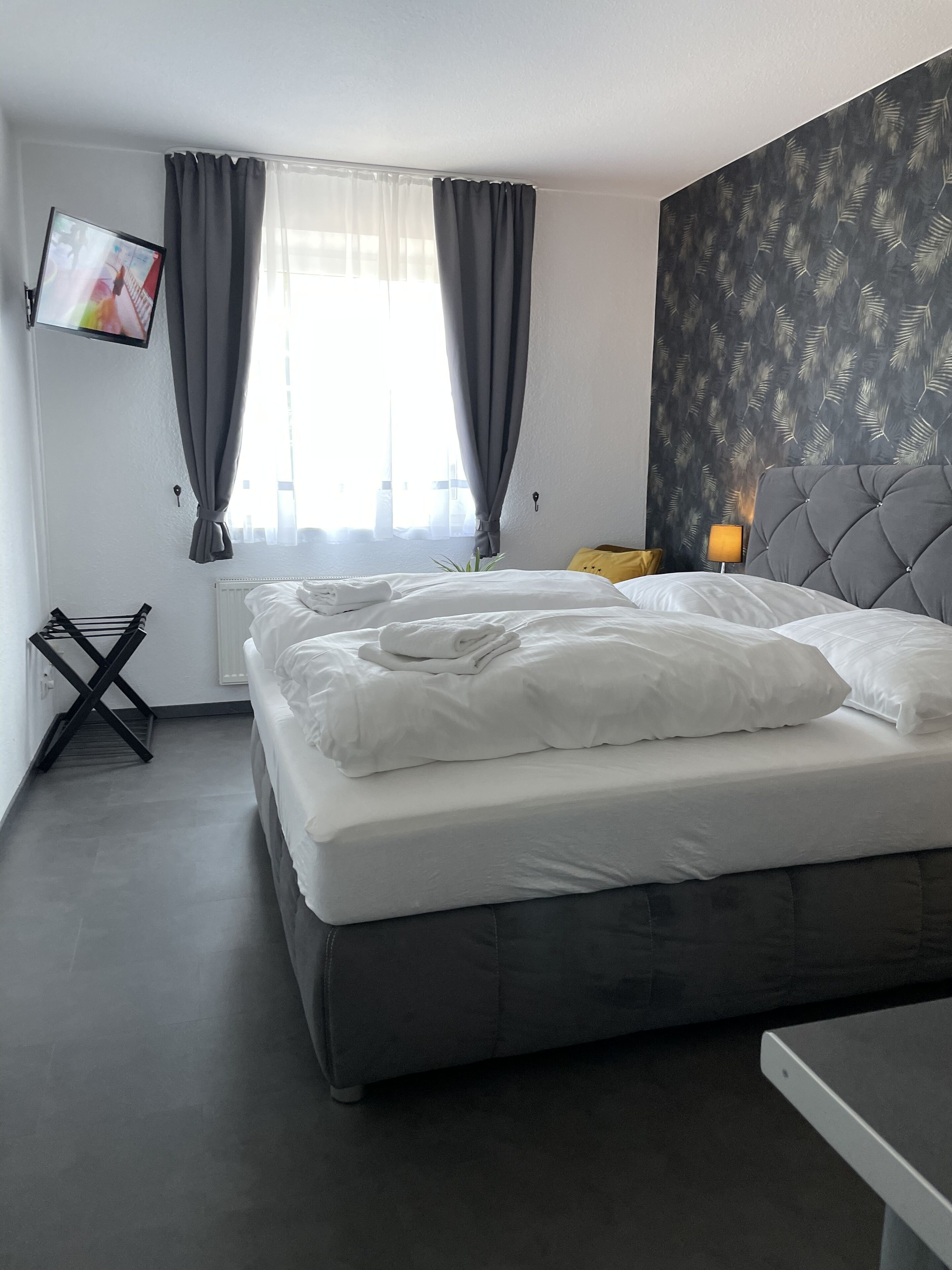 Ein Hotelzimmer mit einem Bett, einem an der Wand befestigten Fernseher, einem Fenster mit Vorhängen und einer Wand mit einem Blattmuster.