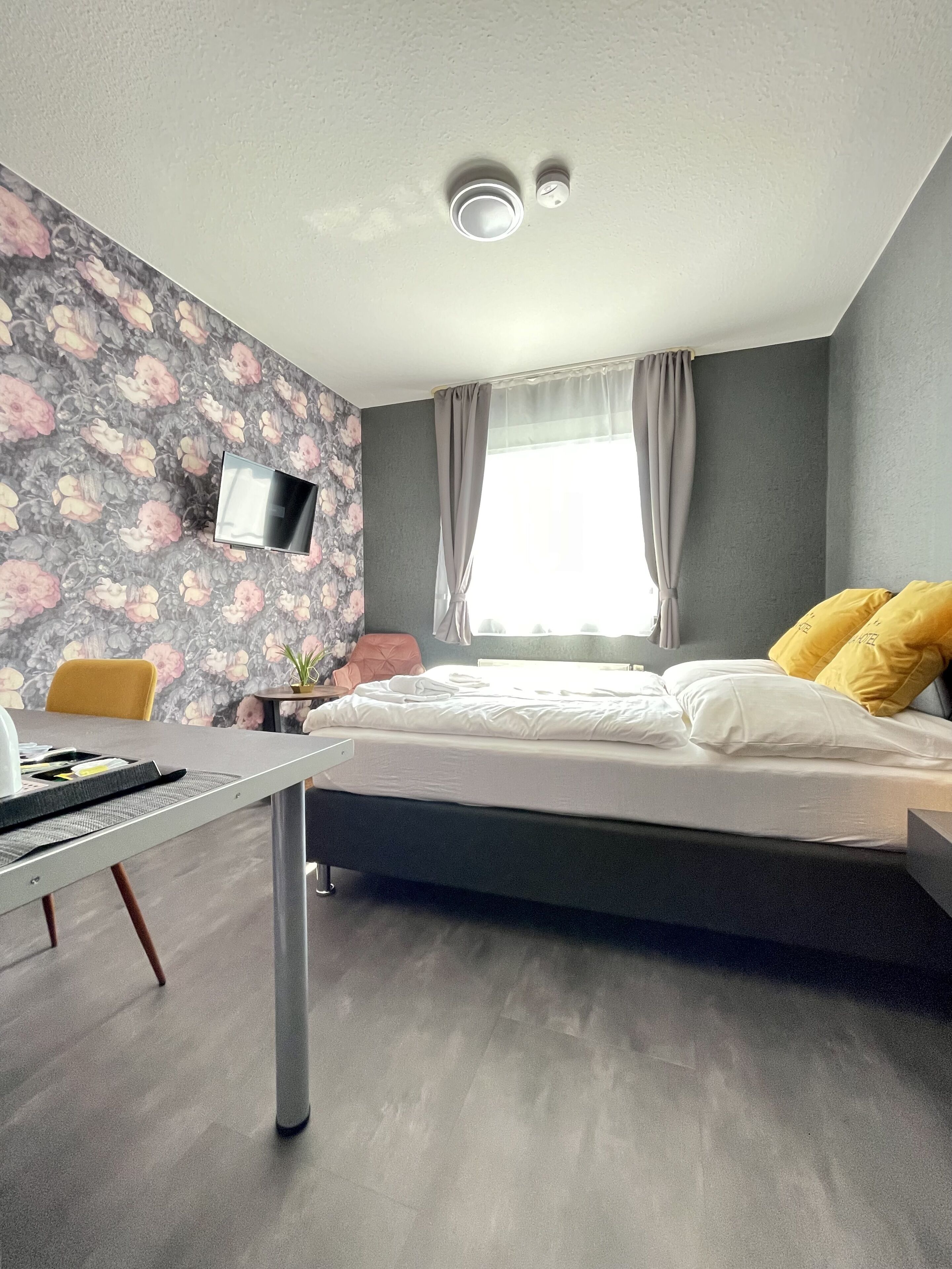 A bedroom with a bed, a desk, a chair, a TV mounted on the wall, and a window with curtains.