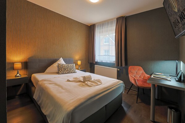 Una habitación de hotel con cama, lámparas de noche, escritorio, silla y radiador.