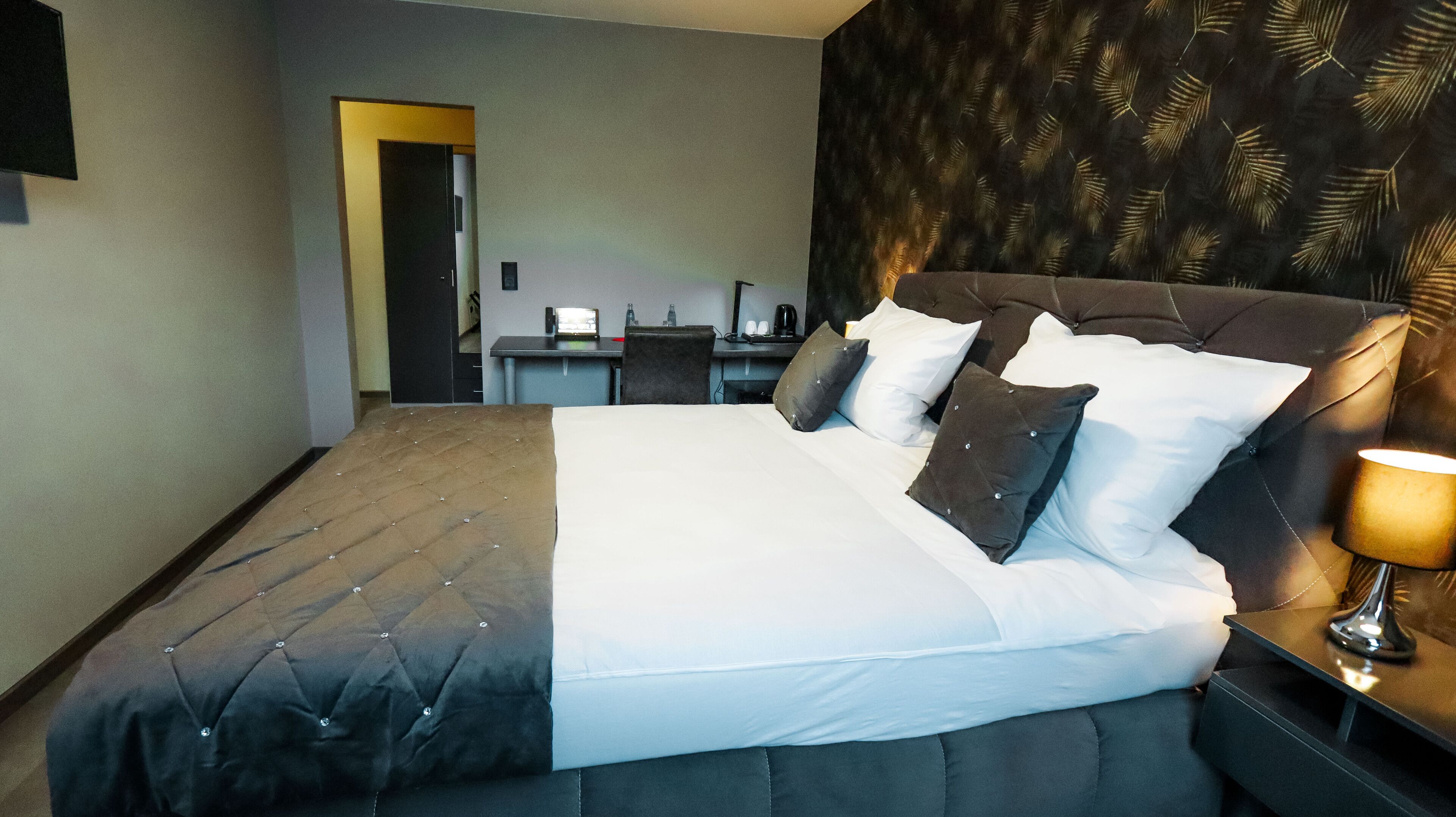 A neatly made bed with a dark headboard and multiple pillows, a bedside table with a lamp, and a small desk with a chair and a mirror.
