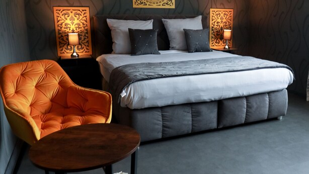 Un dormitorio moderno con una cama grande, dos mesitas de noche con lámparas, un sillón naranja capitonado y una pequeña mesa de madera.