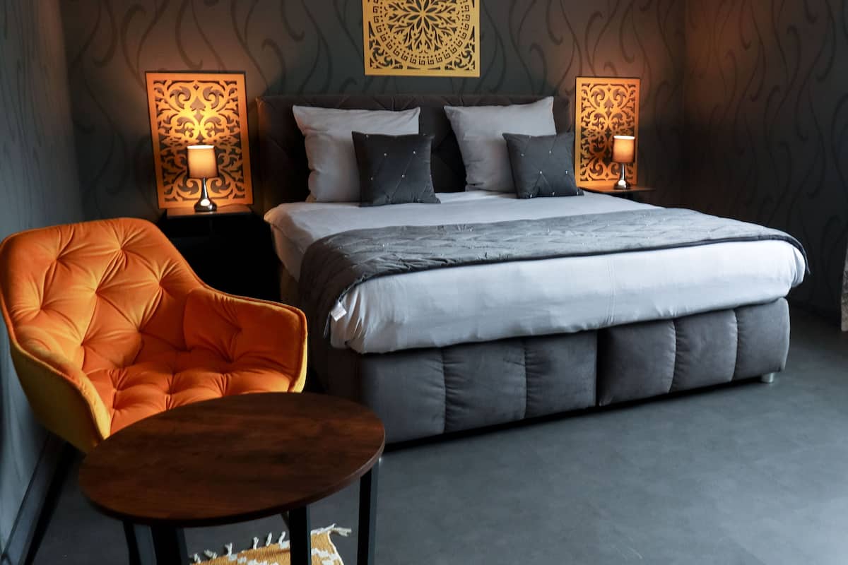 A modern bedroom with a large bed, two bedside tables with lamps, a tufted orange chair, and a small wooden table.