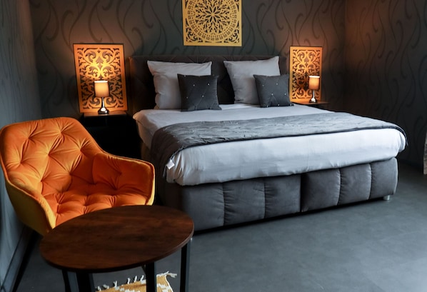 Un dormitorio moderno con una cama grande, dos mesitas de noche con lámparas, un sillón naranja capitonado y una pequeña mesa de madera.