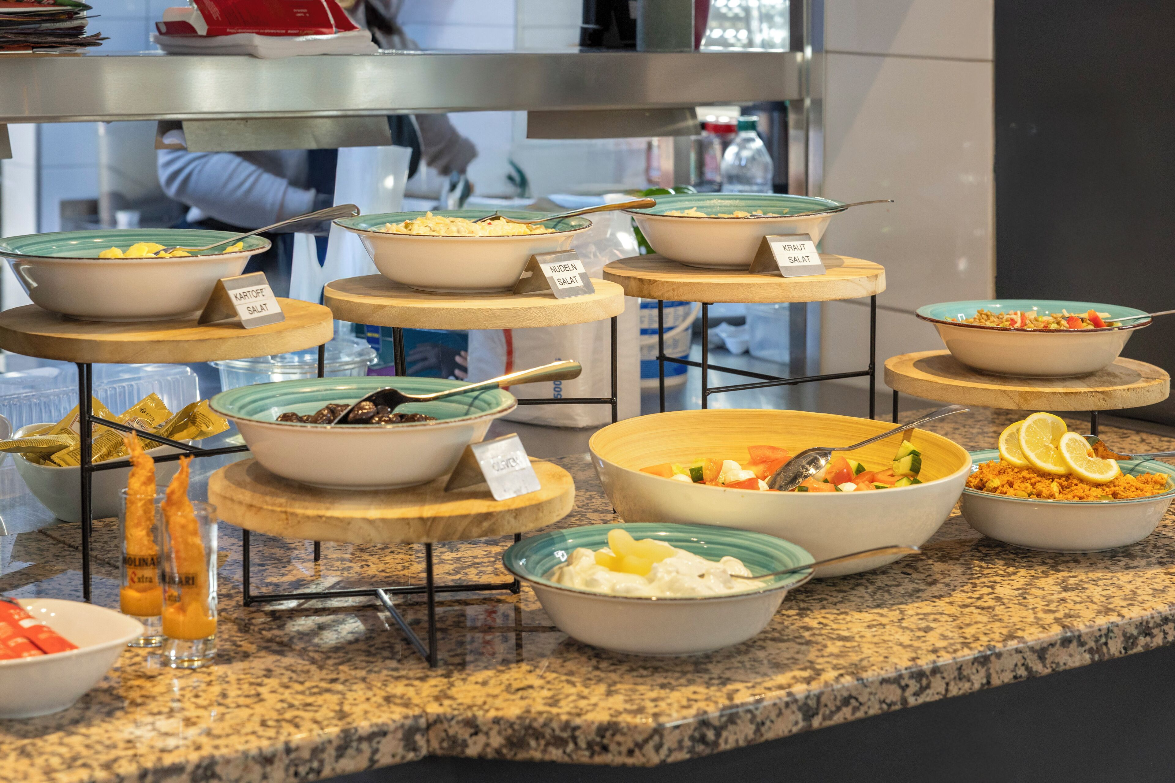 Ein Buffet mit verschiedenen Gerichten, darunter 'NUDEIN SALAT' und 'KARTOFFE SALAT', auf mehreren Etageren angerichtet.