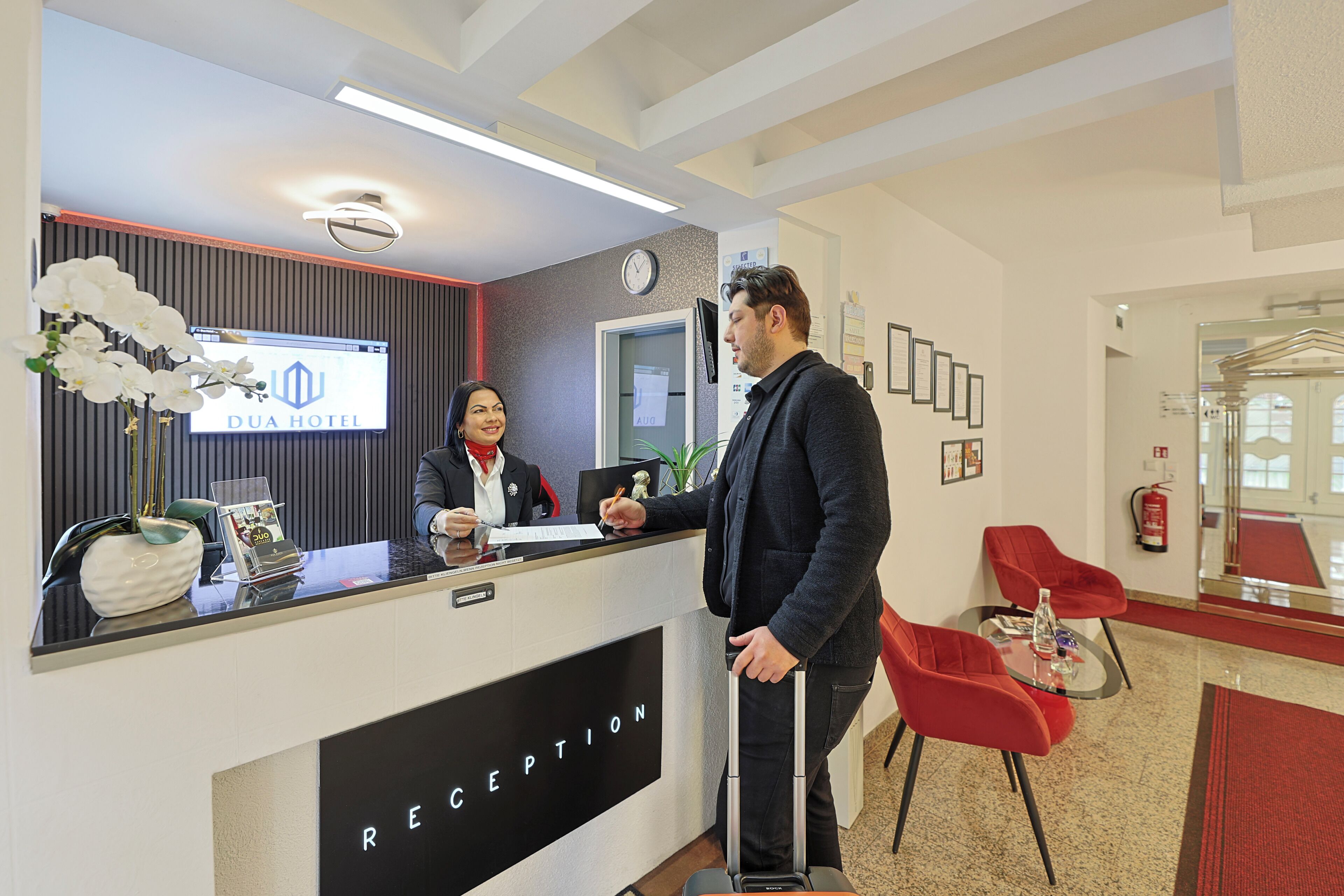 A hotel reception desk with a digital display, reception area, and red chairs.