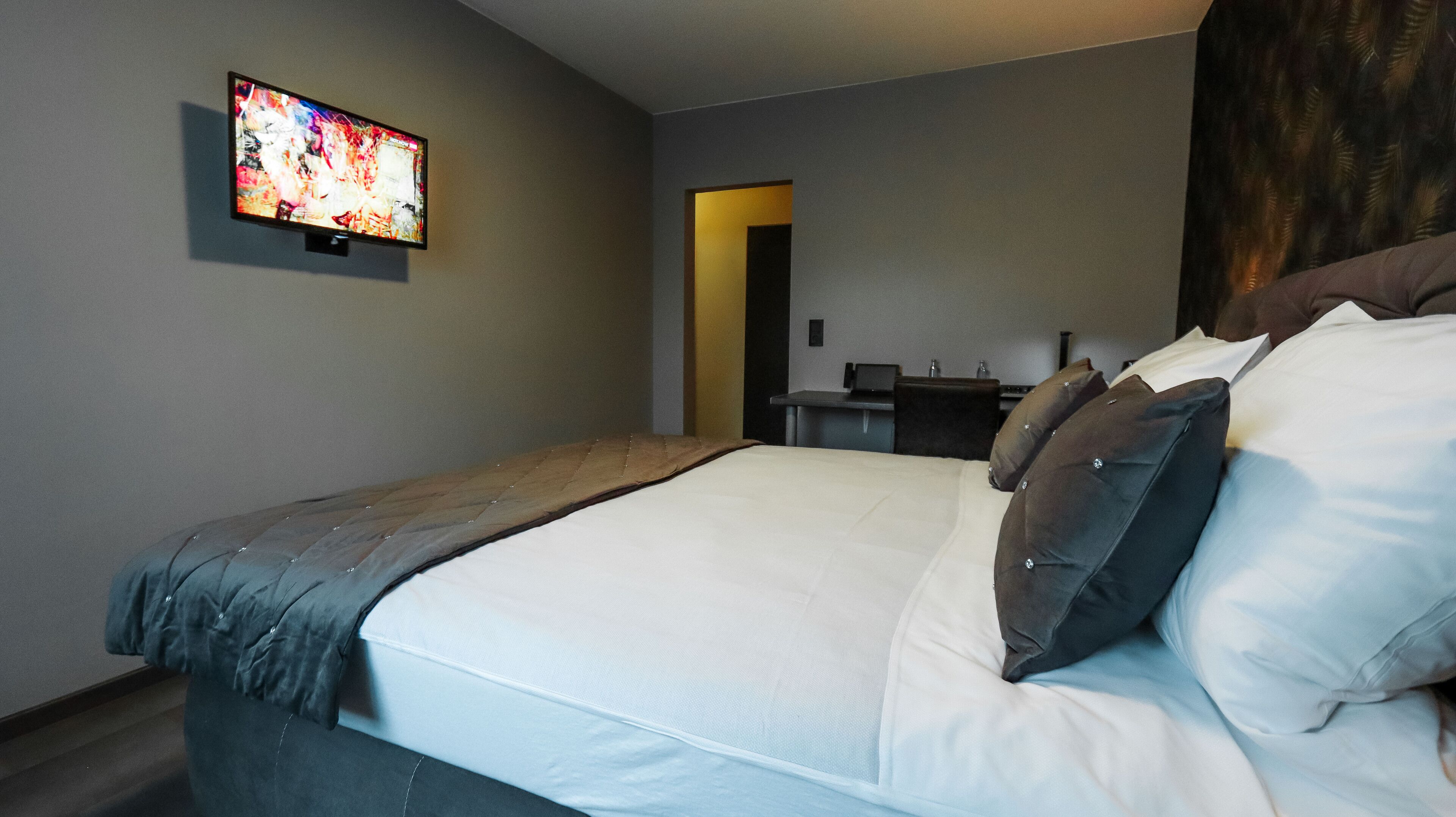 Quarto de hotel moderno com uma cama grande, televisão de tela plana na parede, uma pequena mesa de escritório e uma porta que dá para outro cômodo.