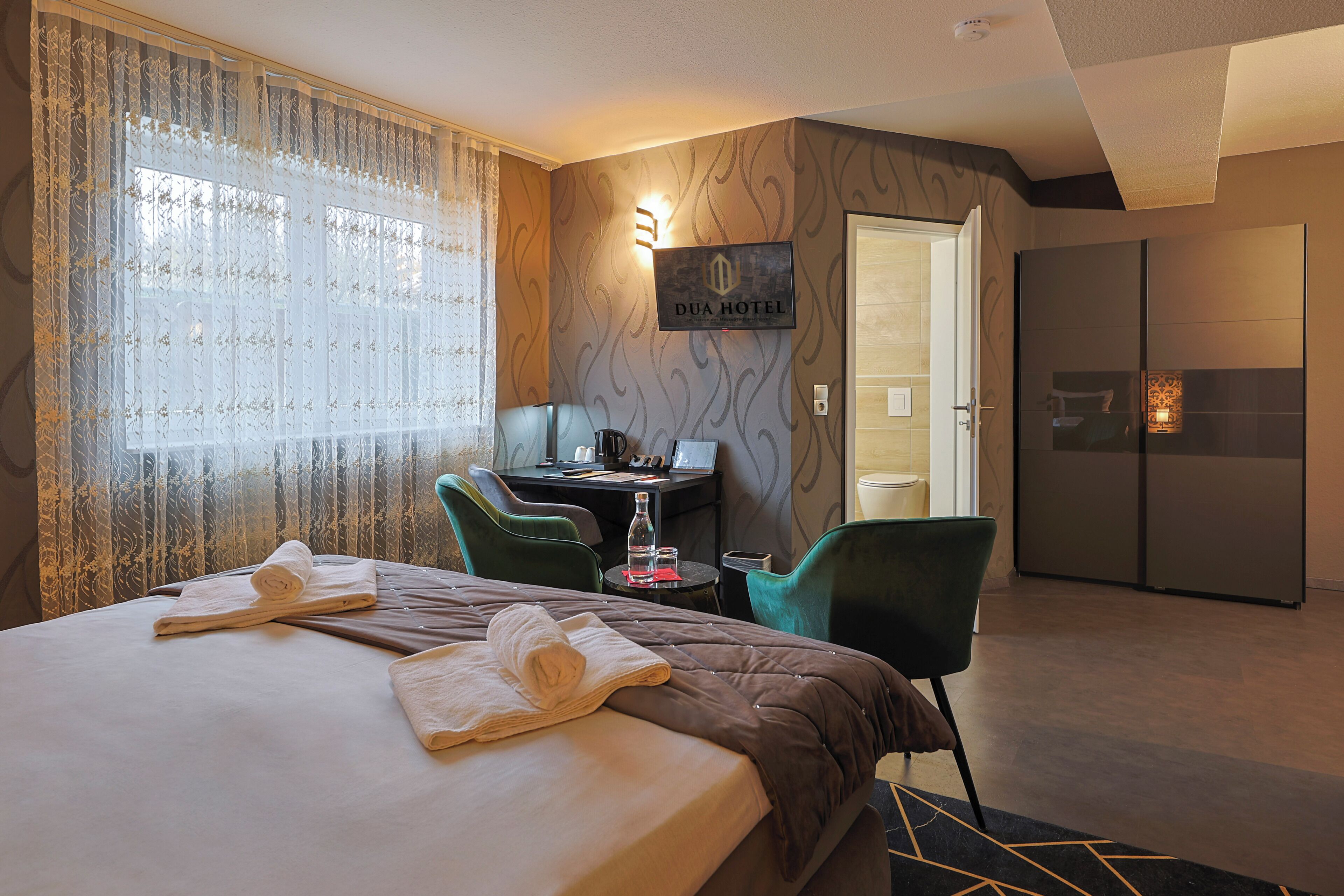 A hotel room with a bed, a desk, a chair, a minibar, and a bathroom visible through an open door.