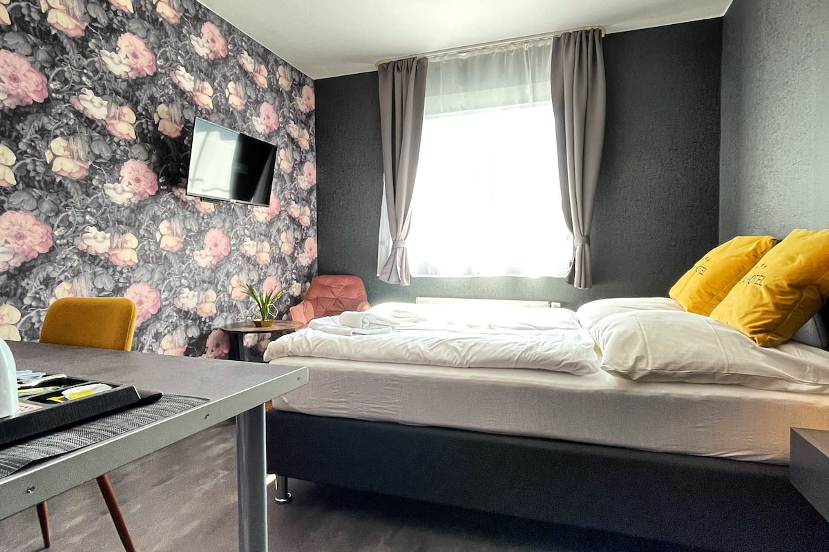 A modern bedroom with a bed, a desk, a chair, a TV, and a window with curtains.