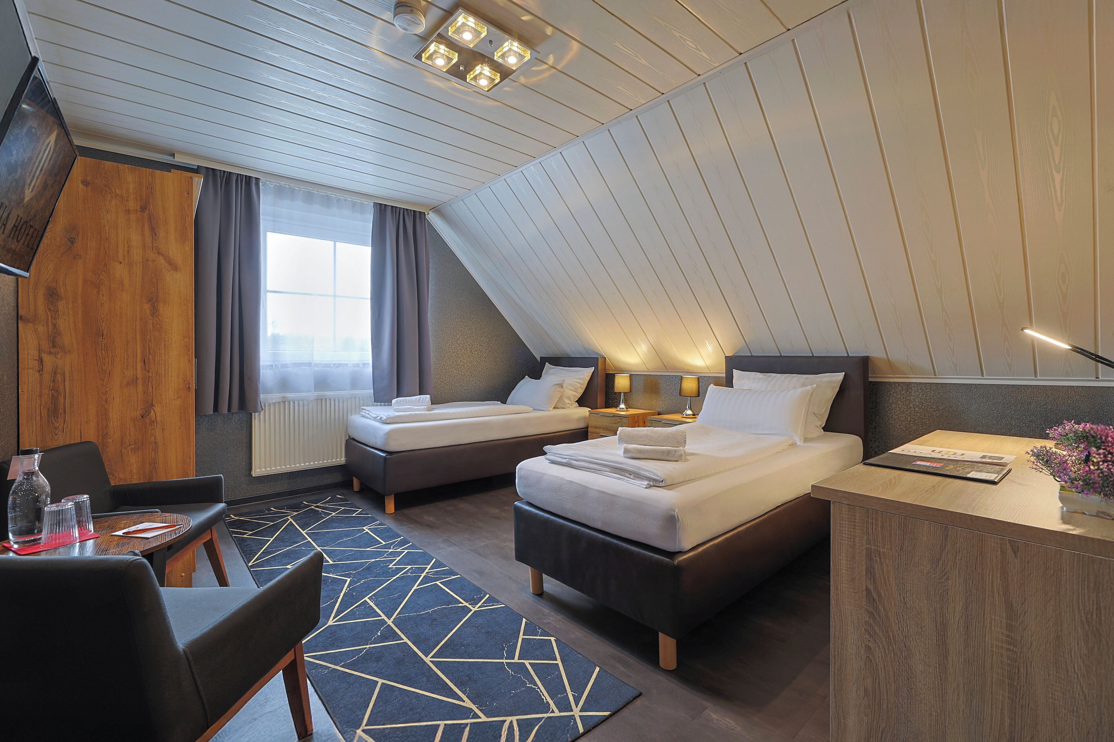 A hotel room with two beds, a desk, a chair, a window with curtains, and a ceiling with recessed lighting.