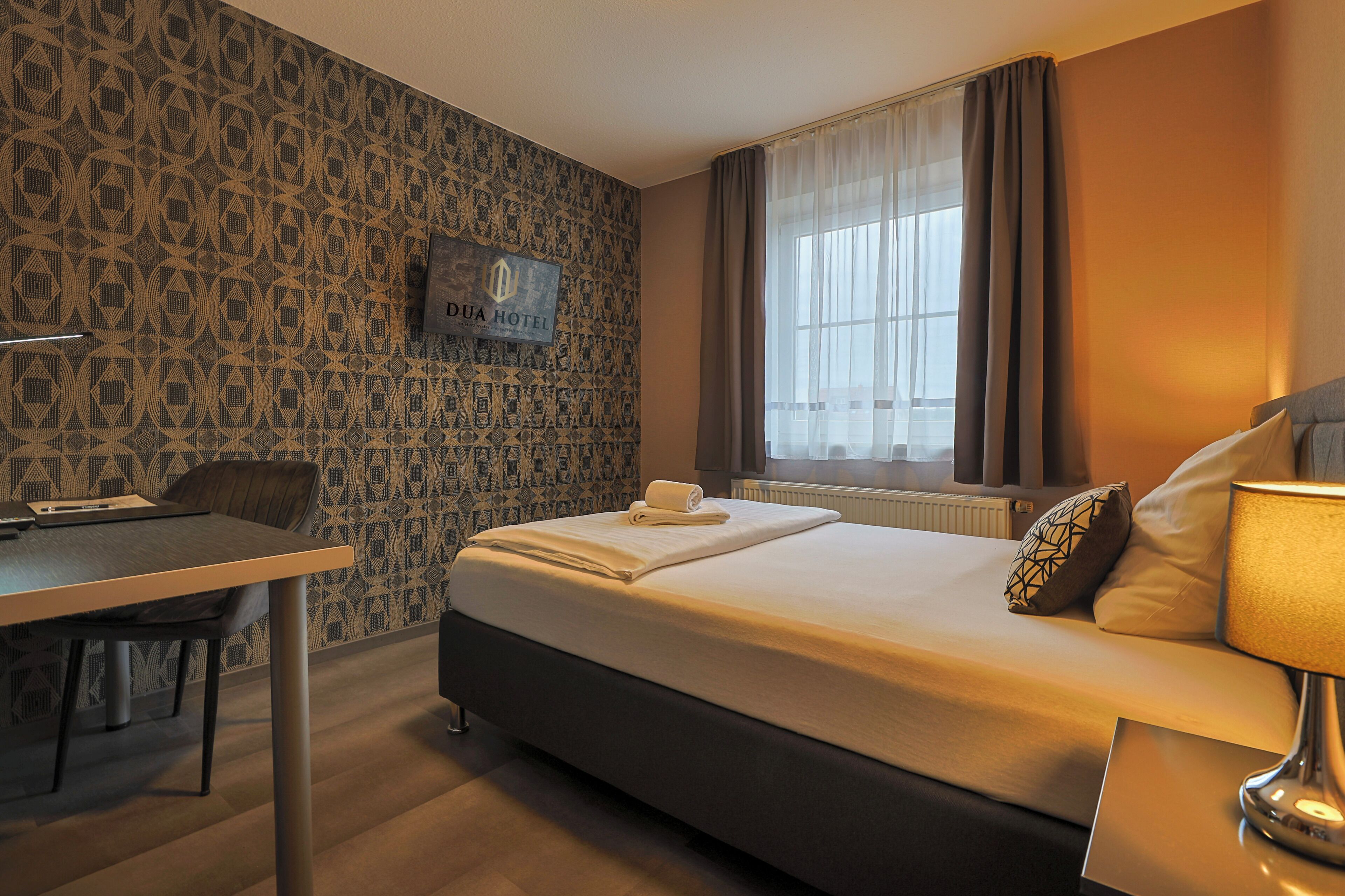 Ein Hotelzimmer mit Bett, Schreibtisch, Stuhl, einem Fenster mit Vorhängen sowie einer Wand mit geometrischem Muster.
