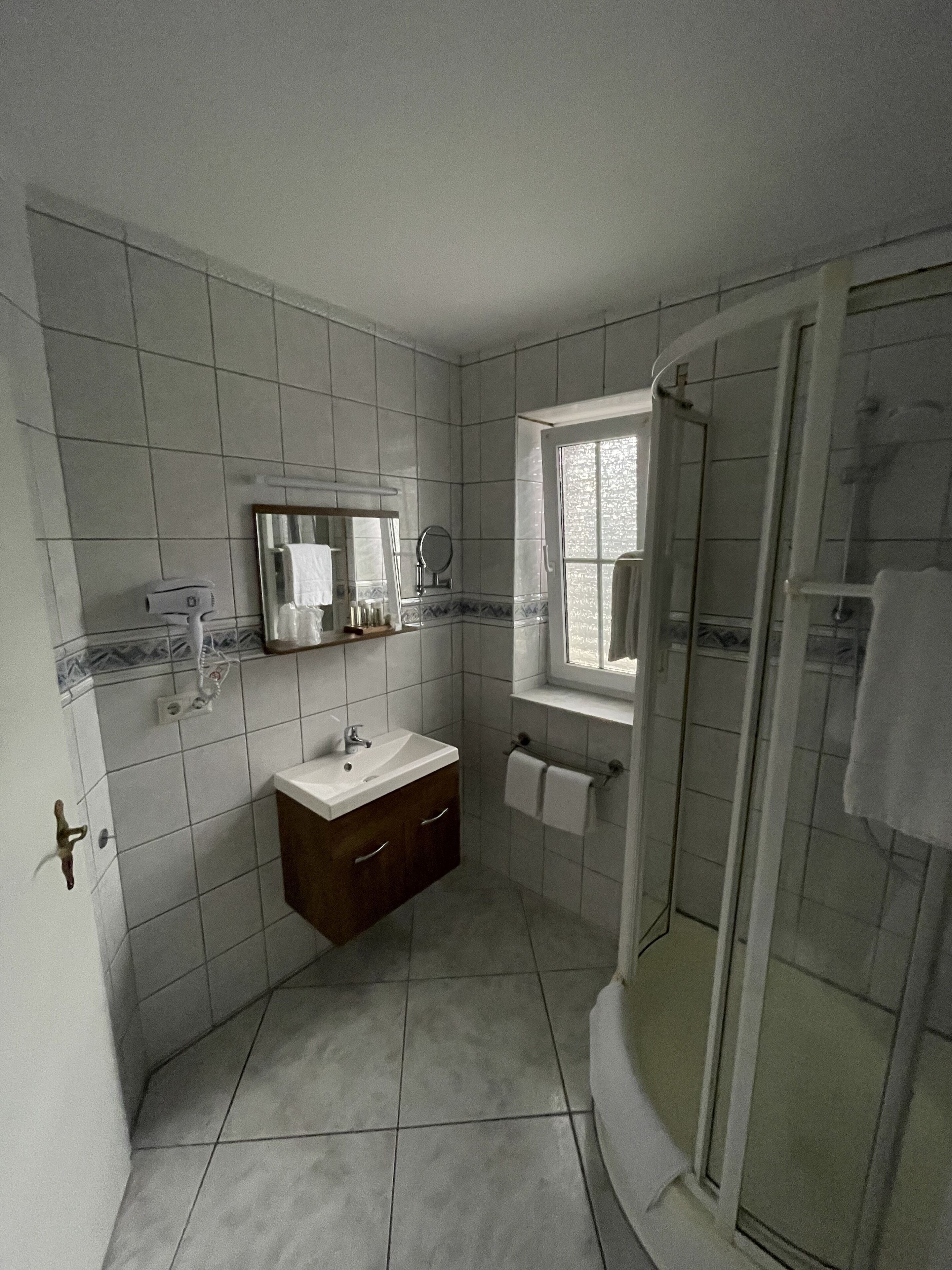 A bathroom with white tiled walls, a sink with a mirror, a window, and a shower area.