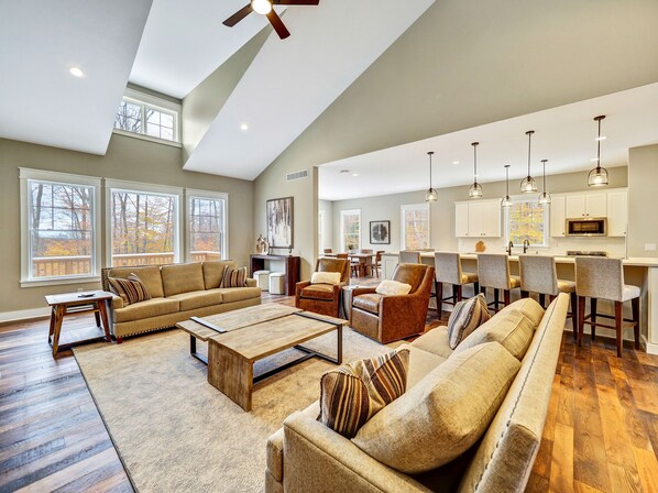 A spacious living room with a large sofa, coffee table, and chairs, a kitchen area with bar stools, and large windows offering views of the outdoors. - Golfers dream w/ 5-star experience! Boyne Mountain ski in/out
Garage, fire-pit! (Boyne Falls)